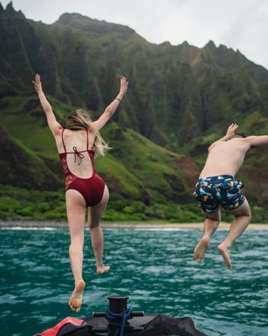 Two people jumping off a boat into turquoise water with Na Pali cliffs in the background, Kauai