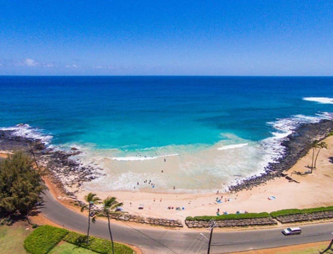 Aerial view of sandy beach cove with turquoise water, rocky outcrops, palm trees, and parked vehicles along coastal road